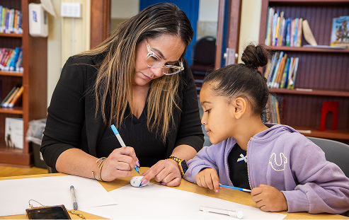 An adult woman with glasses leaning over a desk to assist a young girl with a writing or drawing activity on a piece of paper.