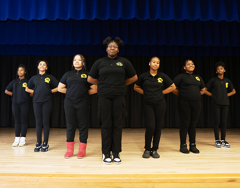 A group of five students dressed in matching black uniforms standing in formation on a stage with blue curtains behind them.
