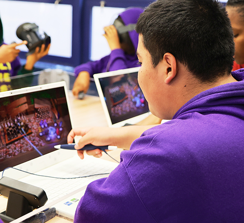 A student in a purple hoodie using a stylus pen on a laptop screen, while another student in the background wears a VR headset