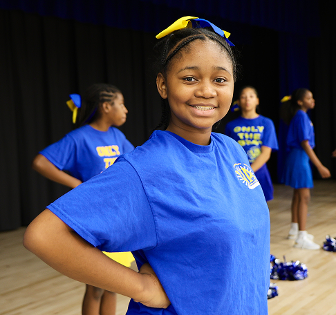 A student in a blue cheerleading outfit smiles while practicing with her team in the school gym.