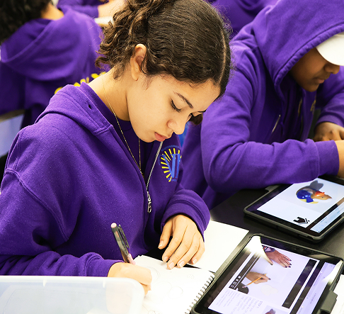 A student with curly hair wearing a purple hoodie, looking down while writing in a notebook next to a tablet showing a video.