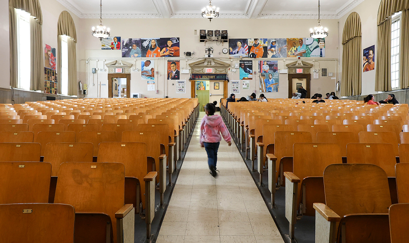 A view from the back of a large school auditorium with rows of wooden seats, showing a student walking down the center aisle toward the front.