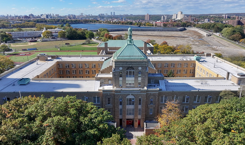 An aerial view of a large, historic brick school building featuring a prominent central tower with a green copper roof, set against a backdrop of trees and a city skyline.