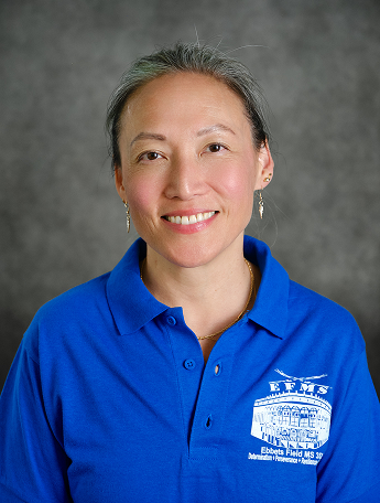 A close-up portrait of a smiling woman with grey-streaked hair tied back, wearing drop earrings and a blue Ebbets Field Middle School polo shirt.