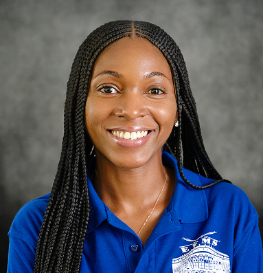 A staff member with long black braided hair smiling warmly, wearing the school's blue uniform polo.