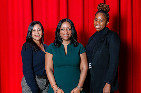 Three professional women standing together against a red curtain background; one in a green dress and two in dark blazers