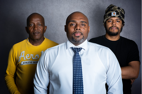 Portrait of three men standing side-by-side, one wearing a yellow t-shirt, other in a white dress shirt and tie, and the last one wearing a black shirt