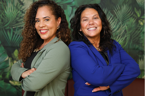 Two professional women smiling while standing back-to-back; one wears a sage green blazer and the other a royal blue blazer