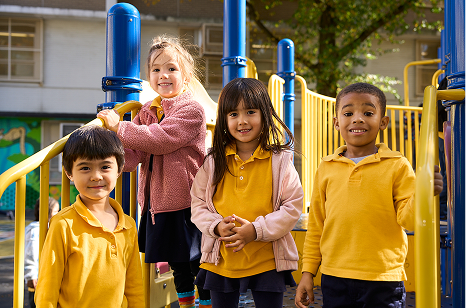 Four young children standing on a playground structure in the sunlight, wearing yellow and pink clothing.