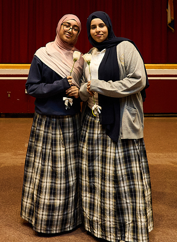 Two female students wearing hijabs and long plaid skirts standing on a stage, holding white roses and smiling