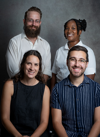 Four smiling staff members posing together against a grey background, including two men with a beard, a woman with braids, and a woman with a black shirt