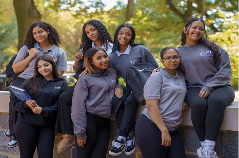 A group of female students wearing matching grey polo shirts posing outdoors on a stone wall