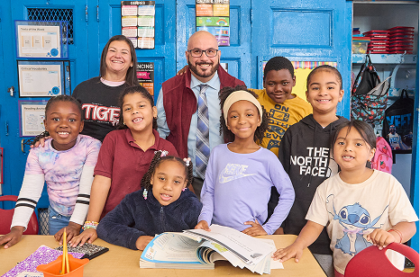 A cheerful group photo inside a classroom featuring two smiling teachers posing with five happy students