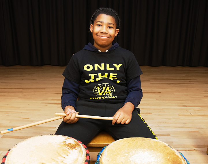 A smiling boy sitting behind a set of drums, holding drumsticks and wearing a black hoodie with yellow text