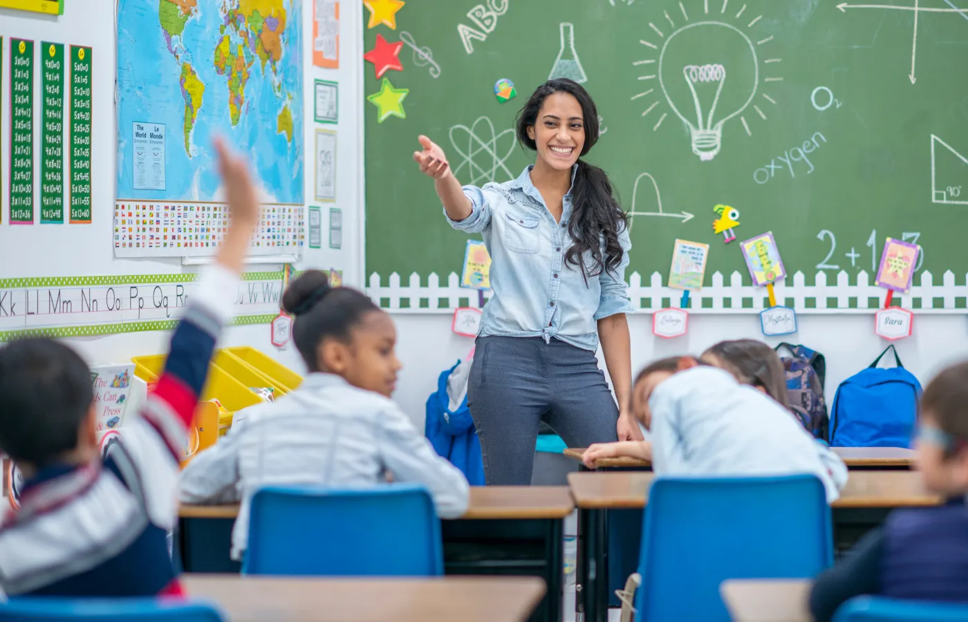 A smiling teacher in a classroom pointing toward a student raising their hand in the back of the room, fostering an engaging and interactive learning environment.