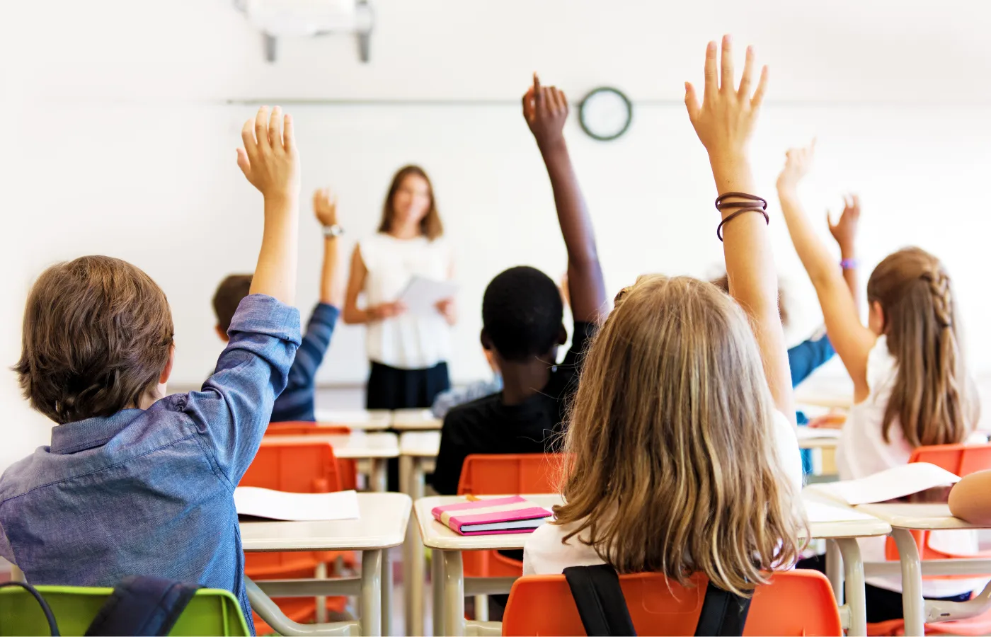Photo taken from the back of a classroom showing students seated at desks with raised hands (back of heads visible), and a blurred teacher at the front of the room, symbolizing effective classroom management and positive behavior.