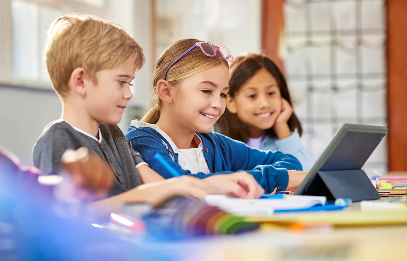 Three students sitting at a classroom table, engaging with an iPad while completing schoolwork, illustrating how Google Classroom supports technology-driven group learning.
