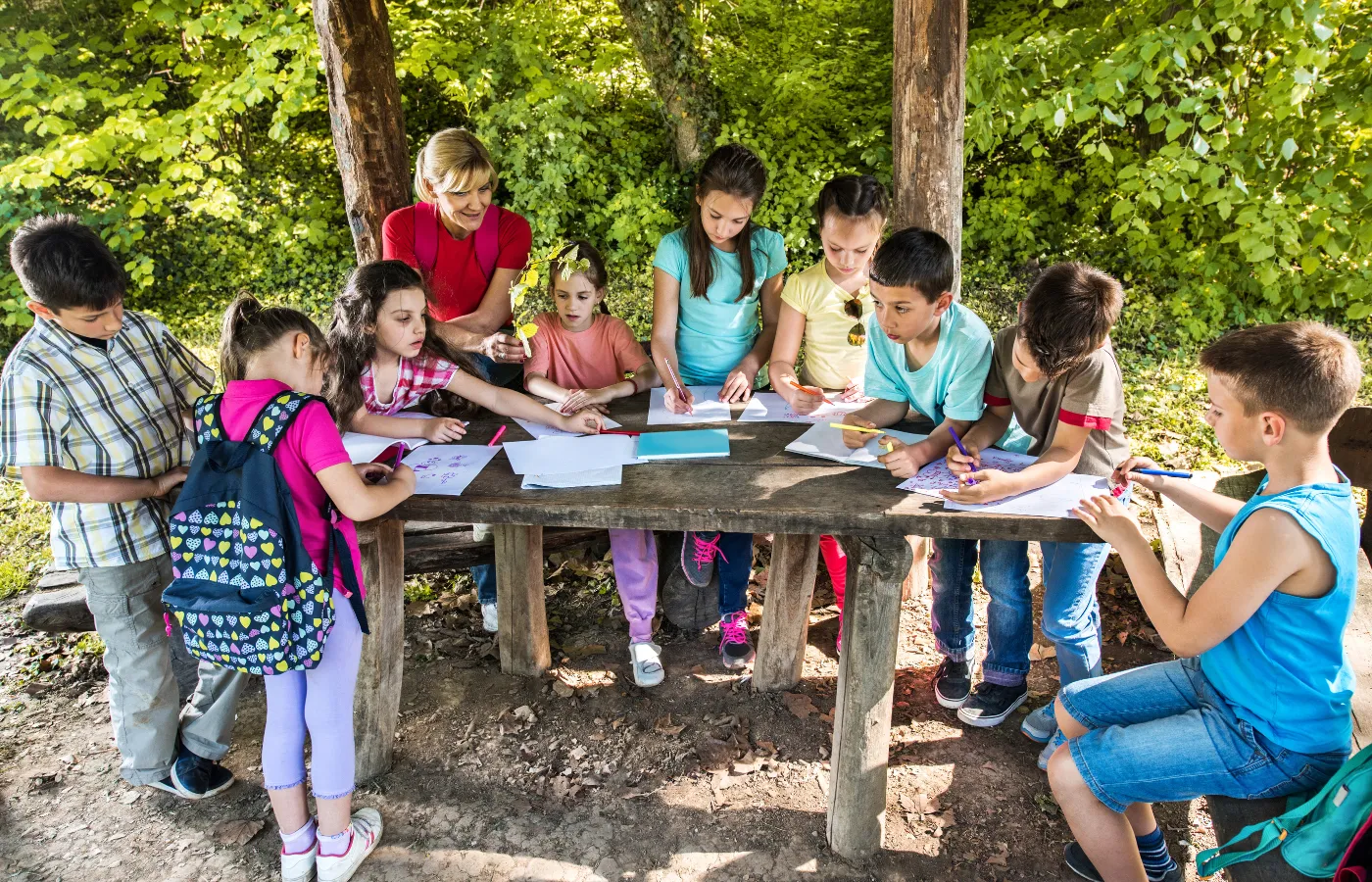 Young students with a teacher working together at a wooden table outdoors, surrounded by nature with a dirt floor and woods in the background. Some students have backpacks, and they are all engaged in a school assignment during a field trip. This image represents a blog idea highlighting a recent school trip in nature.