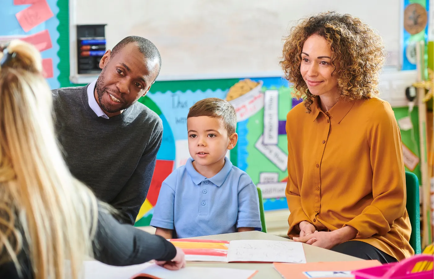 Young child sitting between parents in a classroom, across from a teacher pointing to a book of the student's work, with colorful classroom decorations in the background, illustrating a parent-teacher conference and highlighting the benefits of family engagement in education.