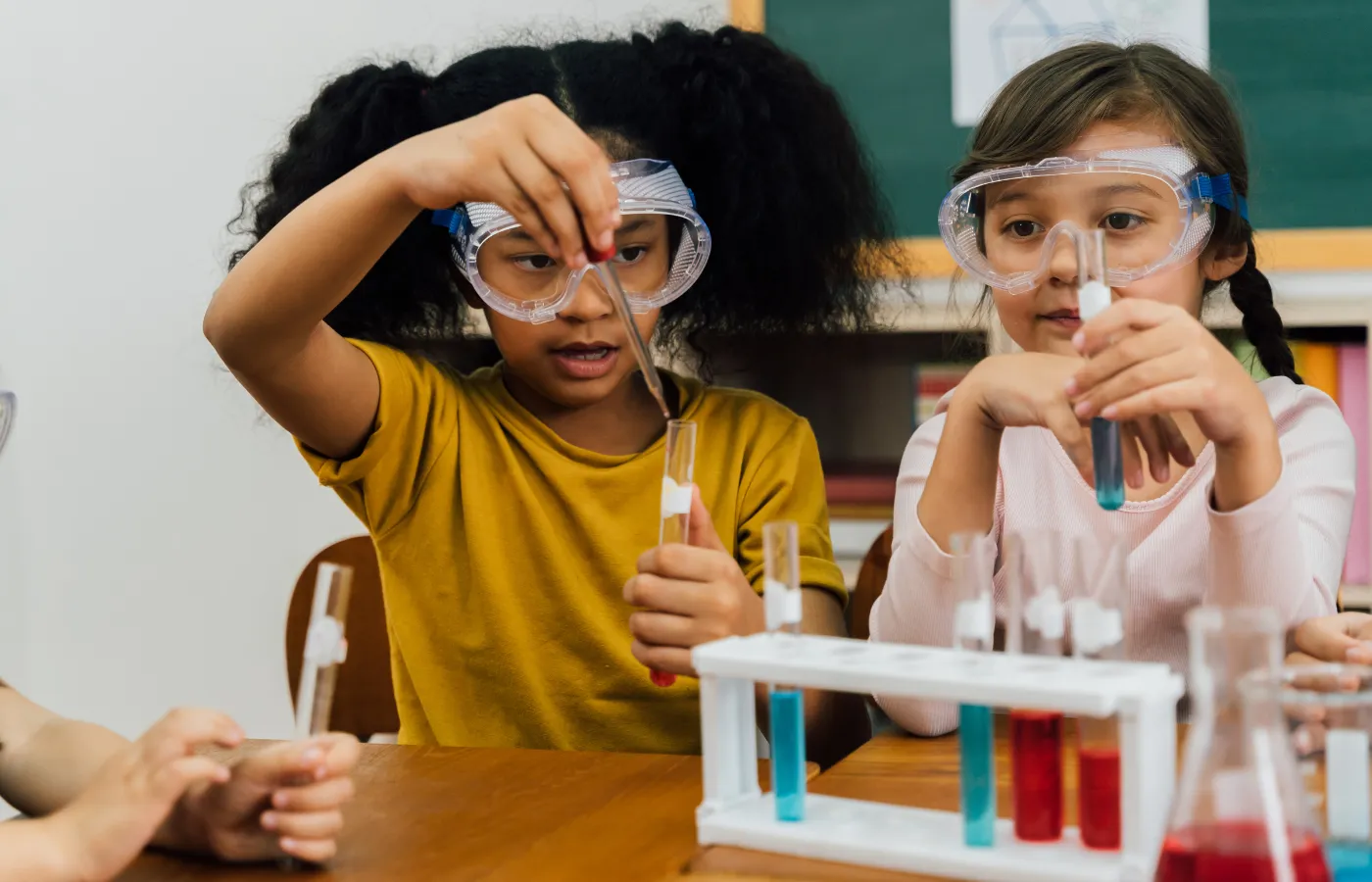 Two students in a science class wearing protective goggles, carefully filling test tubes with red and blue liquid, showcasing a hands-on experiment as an example of a virtual school day activity for prospective parents to observe school day experiences.