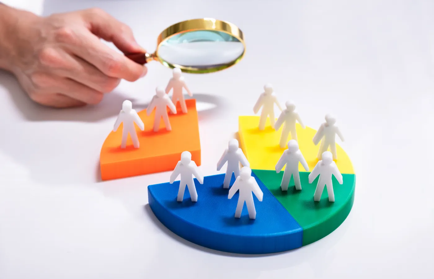 Plastic human figurines placed on a vibrant, multi-colored pie chart, symbolizing audience segmentation, with a hand holding a magnifying glass closely examining the arrangement.