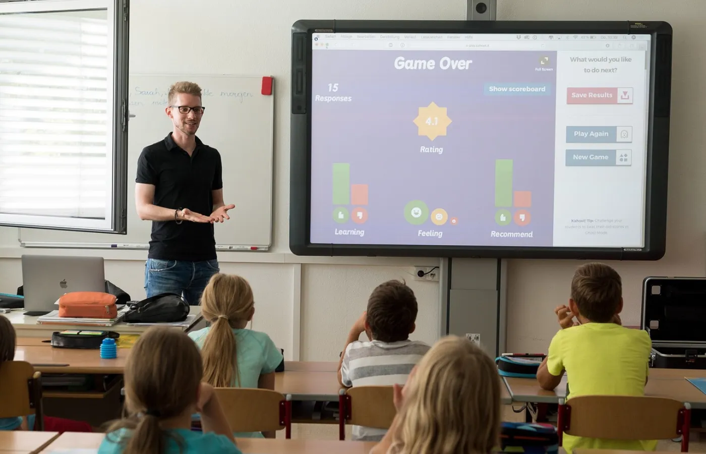 A teacher at the front of a classroom with students seated at desks, looking at a smartboard displaying an interactive learning game, showcasing how Google Classroom integrates third-party tools for engaging, interactive lessons.