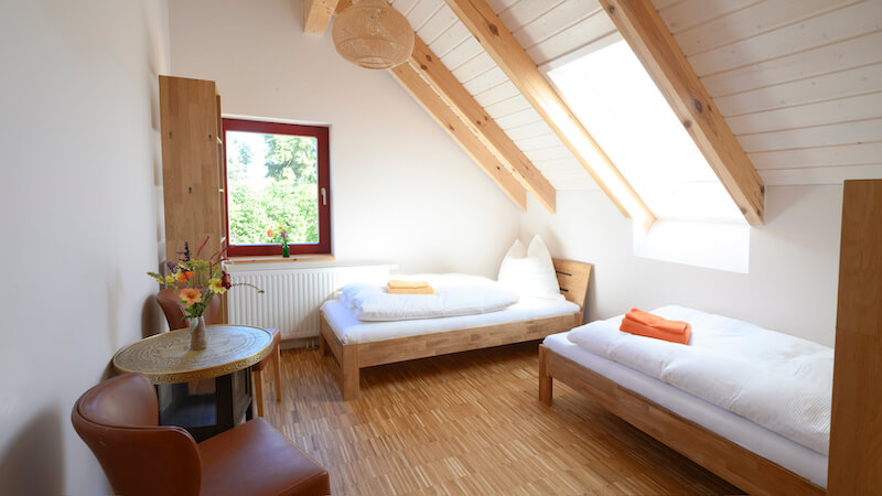 Helles Zweibettzimmer mit Dachschrägen, zwei Einzelbetten mit weißer Bettwäsche und bunten Handtüchern, einem Fenster und einem kleinen runden Tisch mit zwei braunen Stühlen.