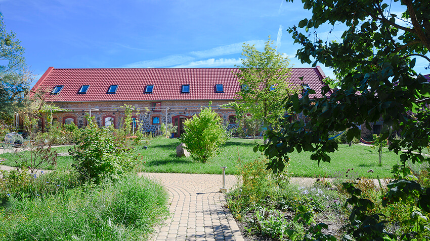 Ländliches Backsteingebäude mit rotem Ziegeldach hinter einem gepflasterten Weg und grünem Garten unter blauem Himmel.