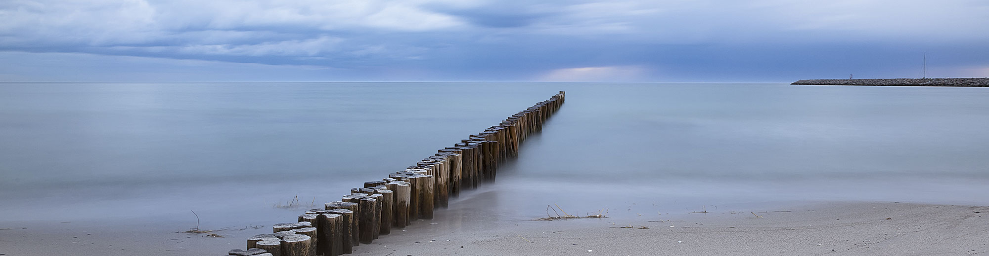 Holzpfähle am ruhigen Strand, die ins Meer und unter bewölktem Himmel führen.