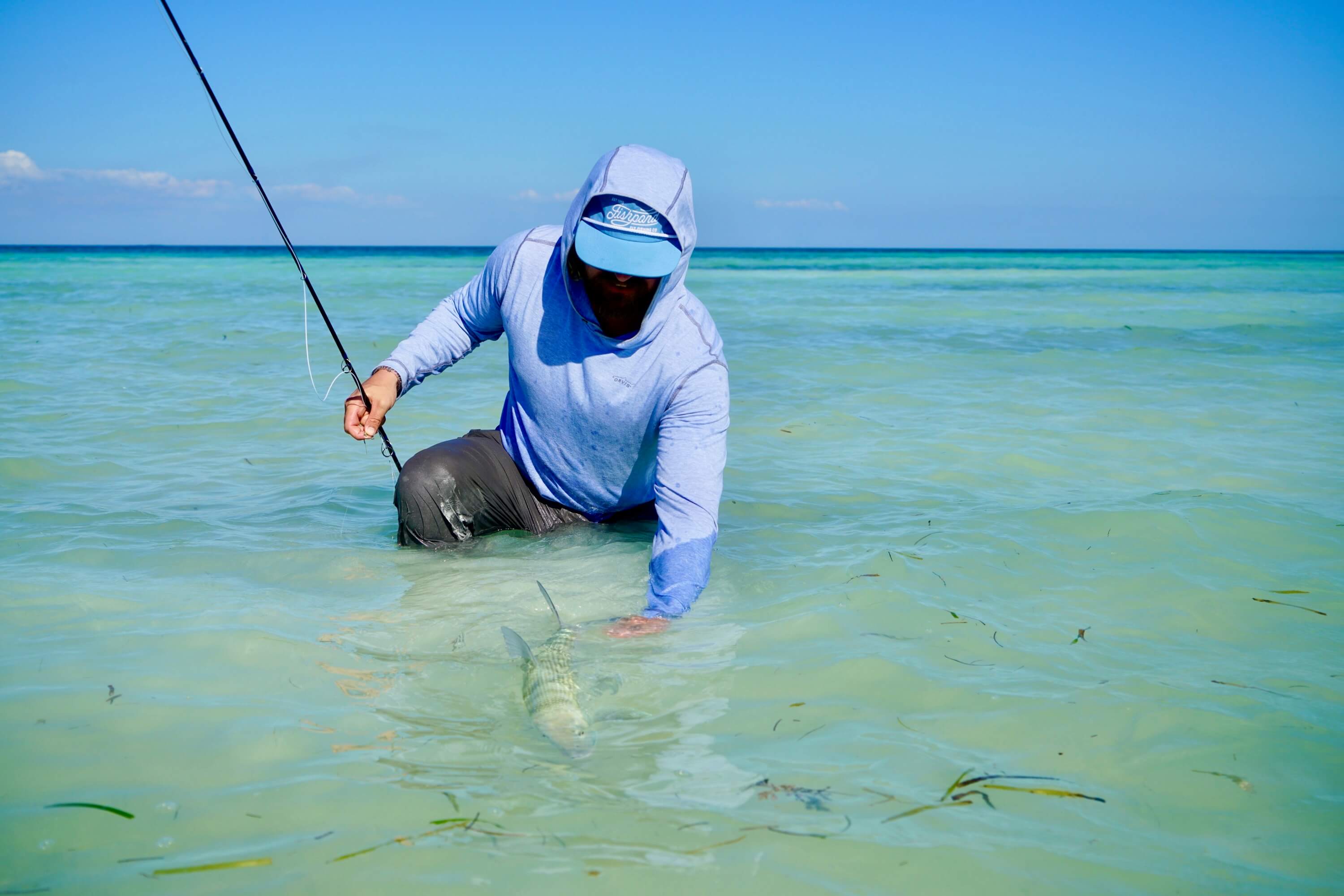 Man in a blue hoodie and cap fishing in shallow clear ocean water and holding a fish.