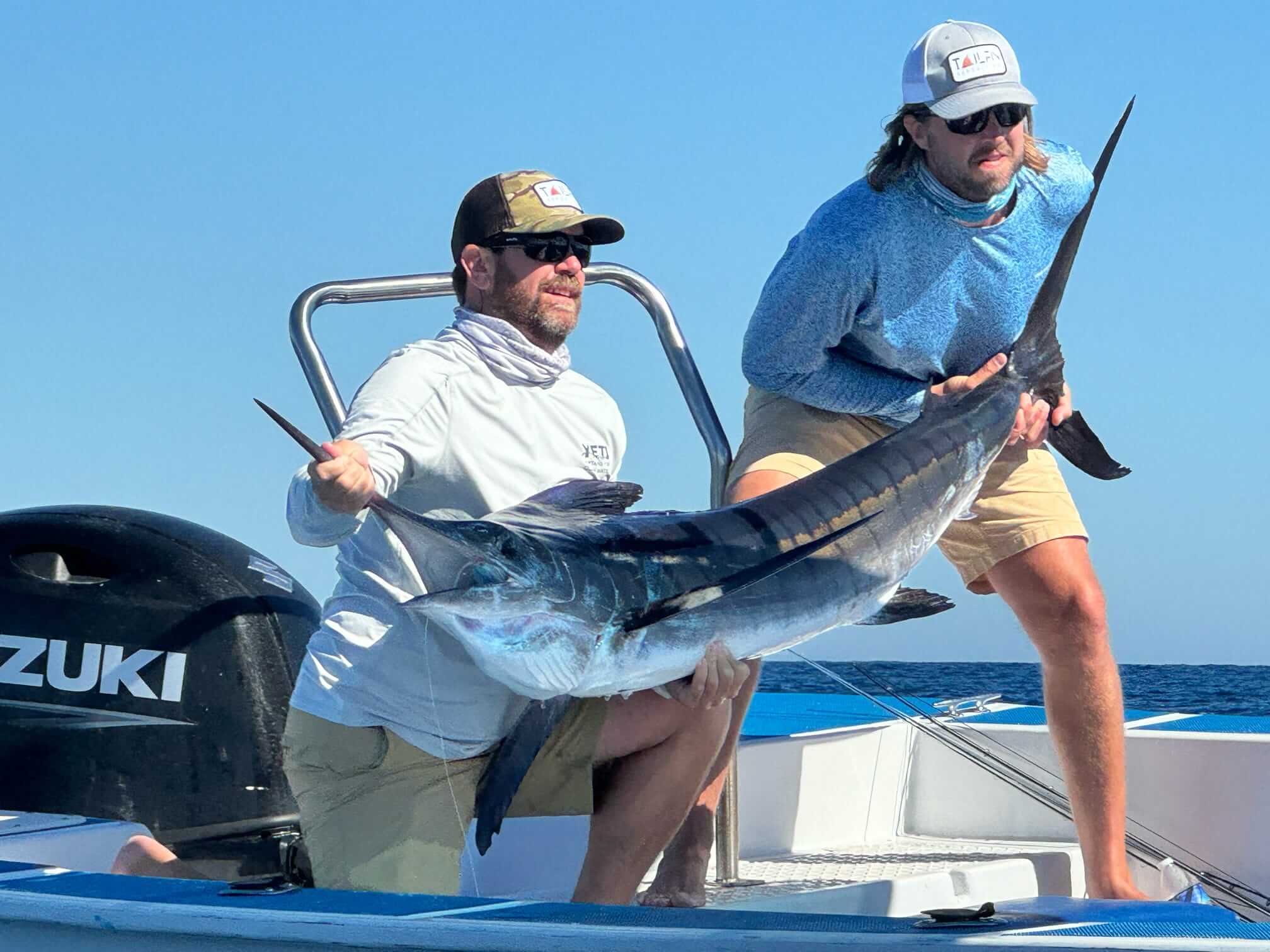 Two men on a boat holding a large marlin with clear blue sky and ocean in the background.