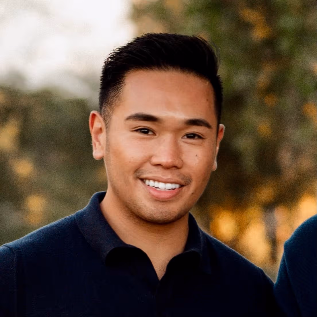 Smiling young man with short black hair wearing a dark collared shirt outdoors with blurred trees in the background.