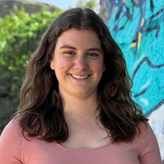 Smiling woman with long brown hair wearing a pink top, standing outdoors near a colorful graffiti wall.