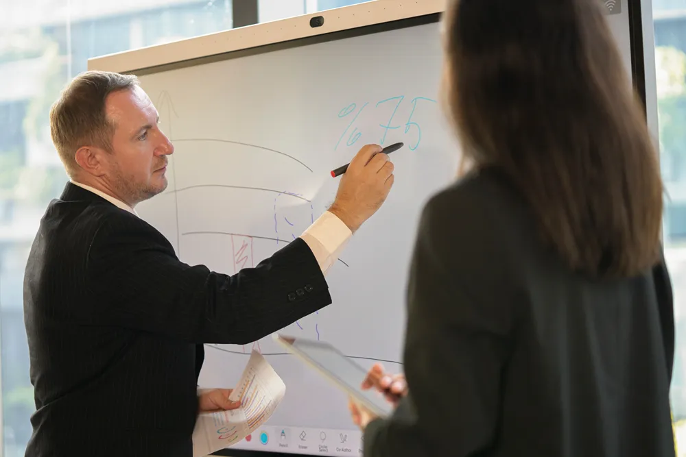 Senior executive mentoring a young colleague using a whiteboard and laptop.