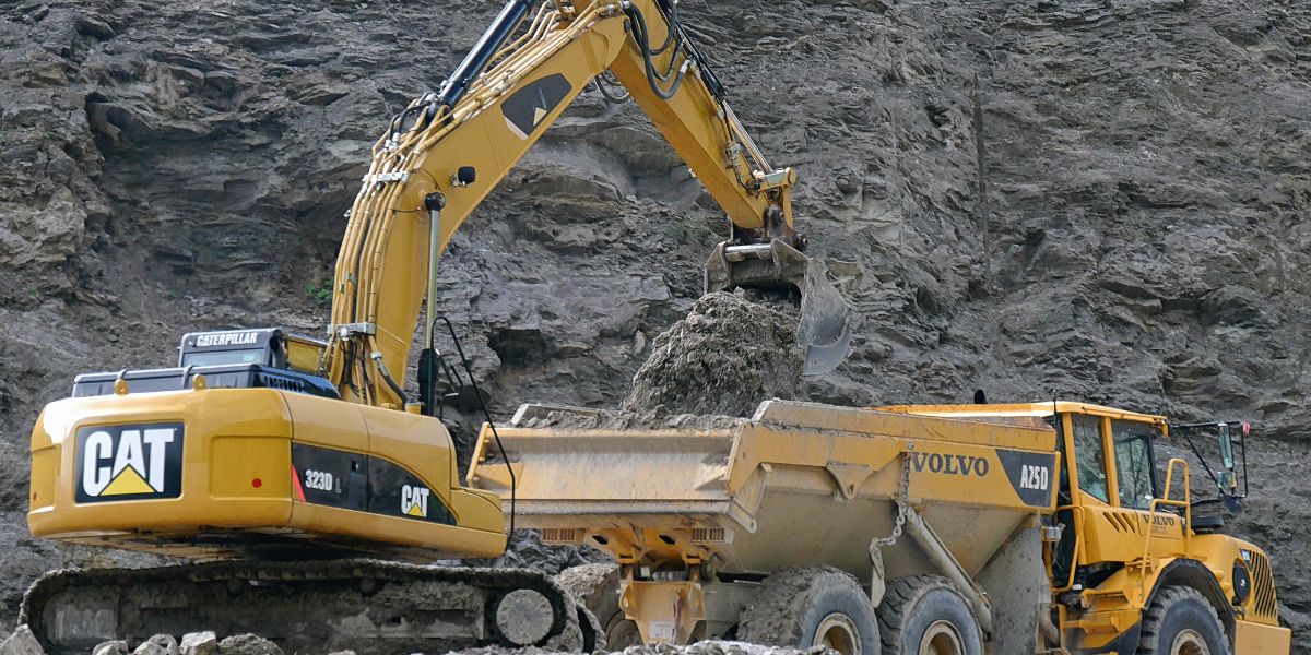 A yellow Caterpillar 323D L excavator loading dirt into the back of a yellow Volvo A25D articulated dump truck at a rocky construction or mining site.