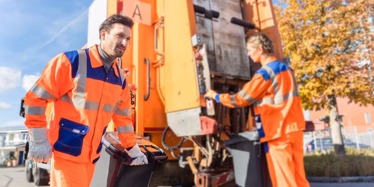 Man wheeling bin on side on refuse vehicle, wearing bright orange Hi Vis Coat, with other worker behind loading bin into back of bin lorry.