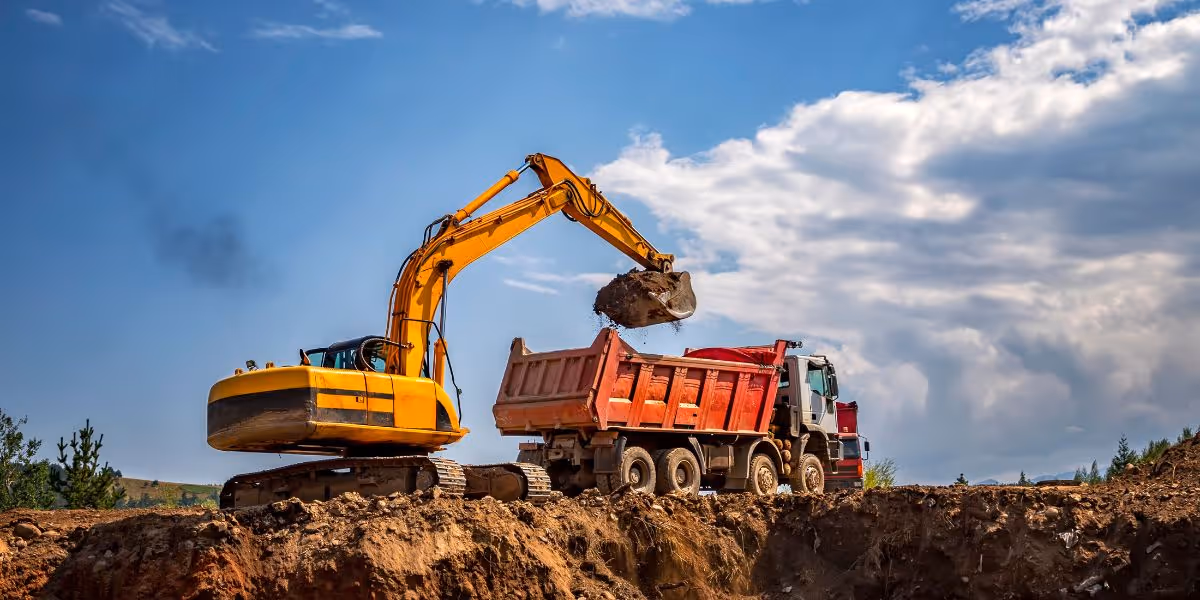 Excavator loading soil into a tipper truck on a construction site under a blue sky, representing heavy equipment and logistics in the construction industry.