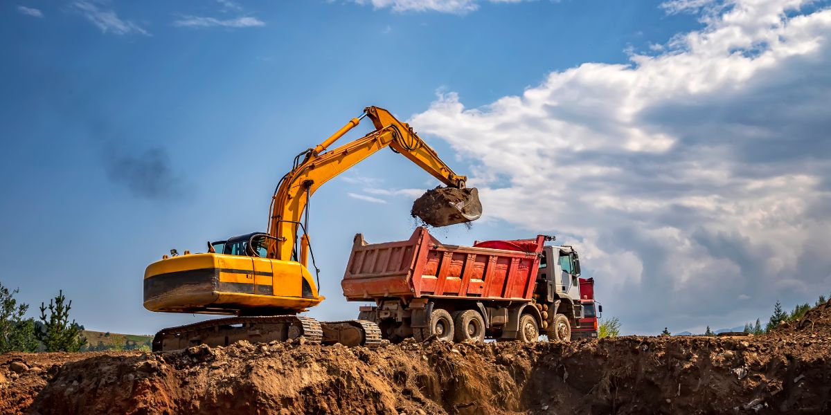 Excavator loading soil into a tipper truck on a construction site under a blue sky, representing heavy equipment and logistics in the construction industry.