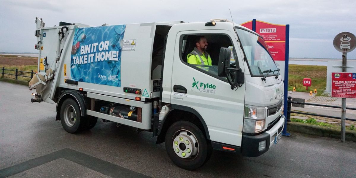 Fylde Council waste collection vehicle parked by the roadside, with a driver in the cab and a large “Bin it or take it home!” campaign graphic displayed on the side.