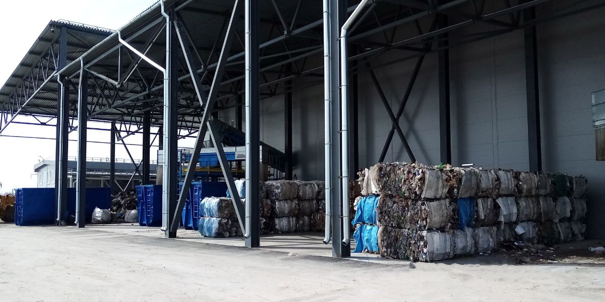 Industrial warehouse interior with metal beams and skylights, showing parked blue equipment on the left and stacked pallets, boxes, and materials on the right.