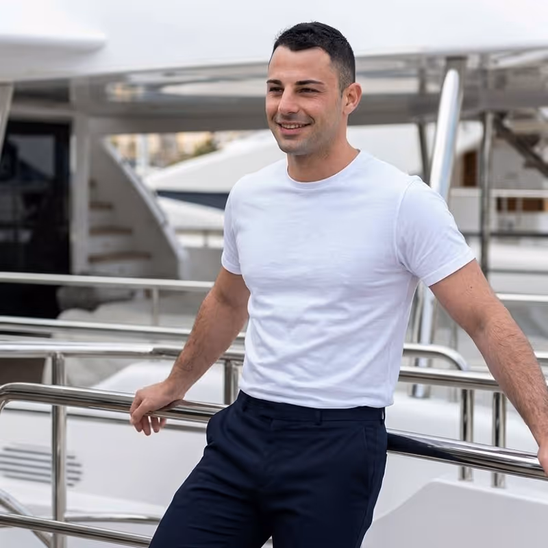Smiling man in a white t-shirt and dark pants leaning on a boat railing on a yacht.