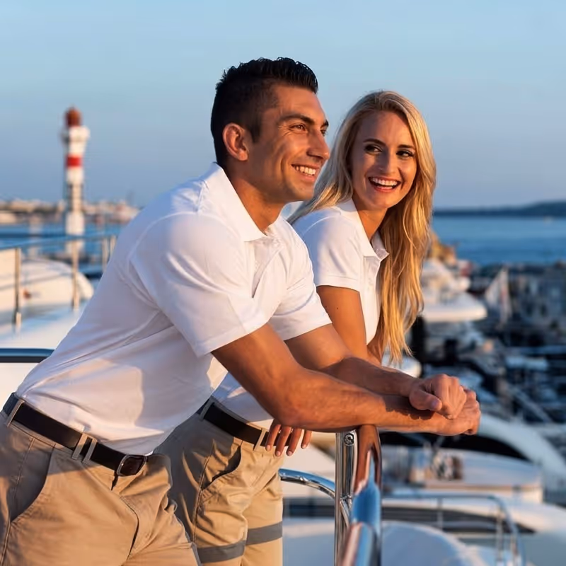 Smiling man and woman in white polo shirts leaning on a yacht railing overlooking a marina at sunset.