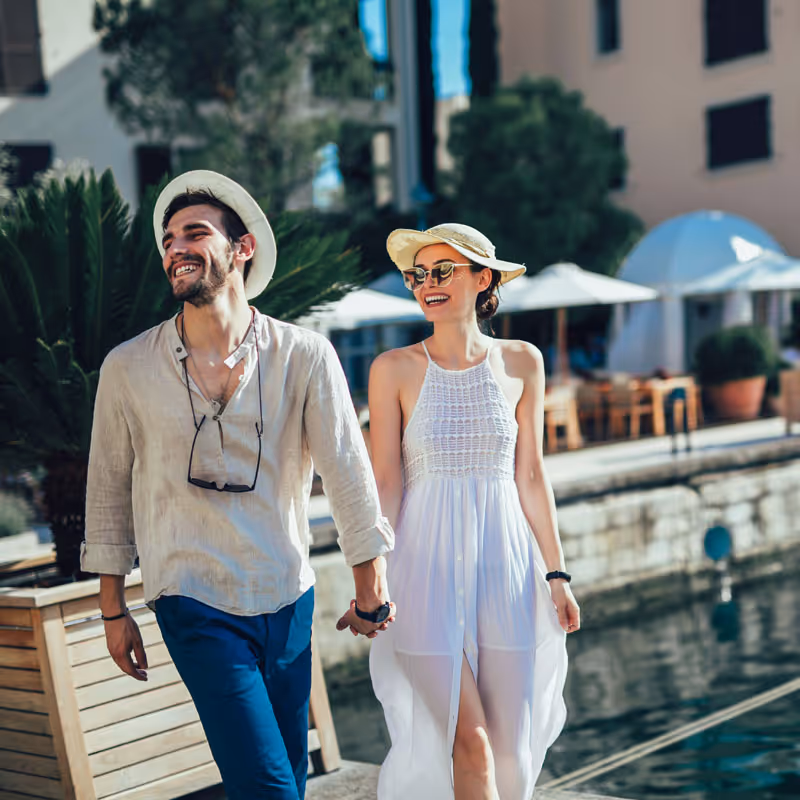 Smiling couple holding hands and walking by a waterfront with outdoor dining in the background on a sunny day.