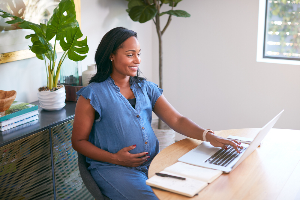 Pregnant woman sitting at a table using a laptop with a notebook and pen nearby.