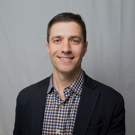 Smiling man with short brown hair wearing a checkered shirt and dark blazer against gray background.