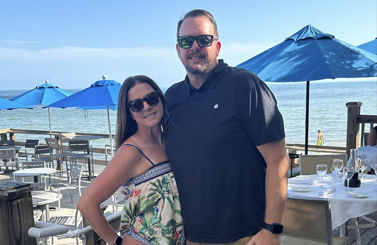 Man and woman wearing sunglasses pose smiling at a seaside restaurant with blue umbrellas and ocean view.
