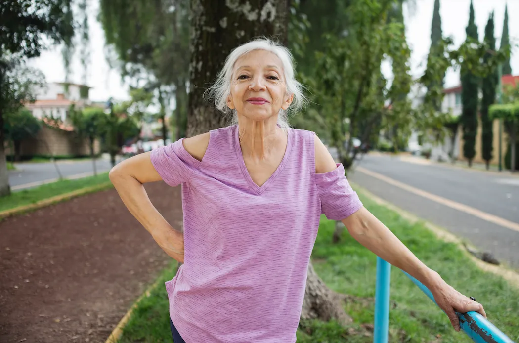 Smiling older woman in a purple top stretching outdoors by a park path and railing.