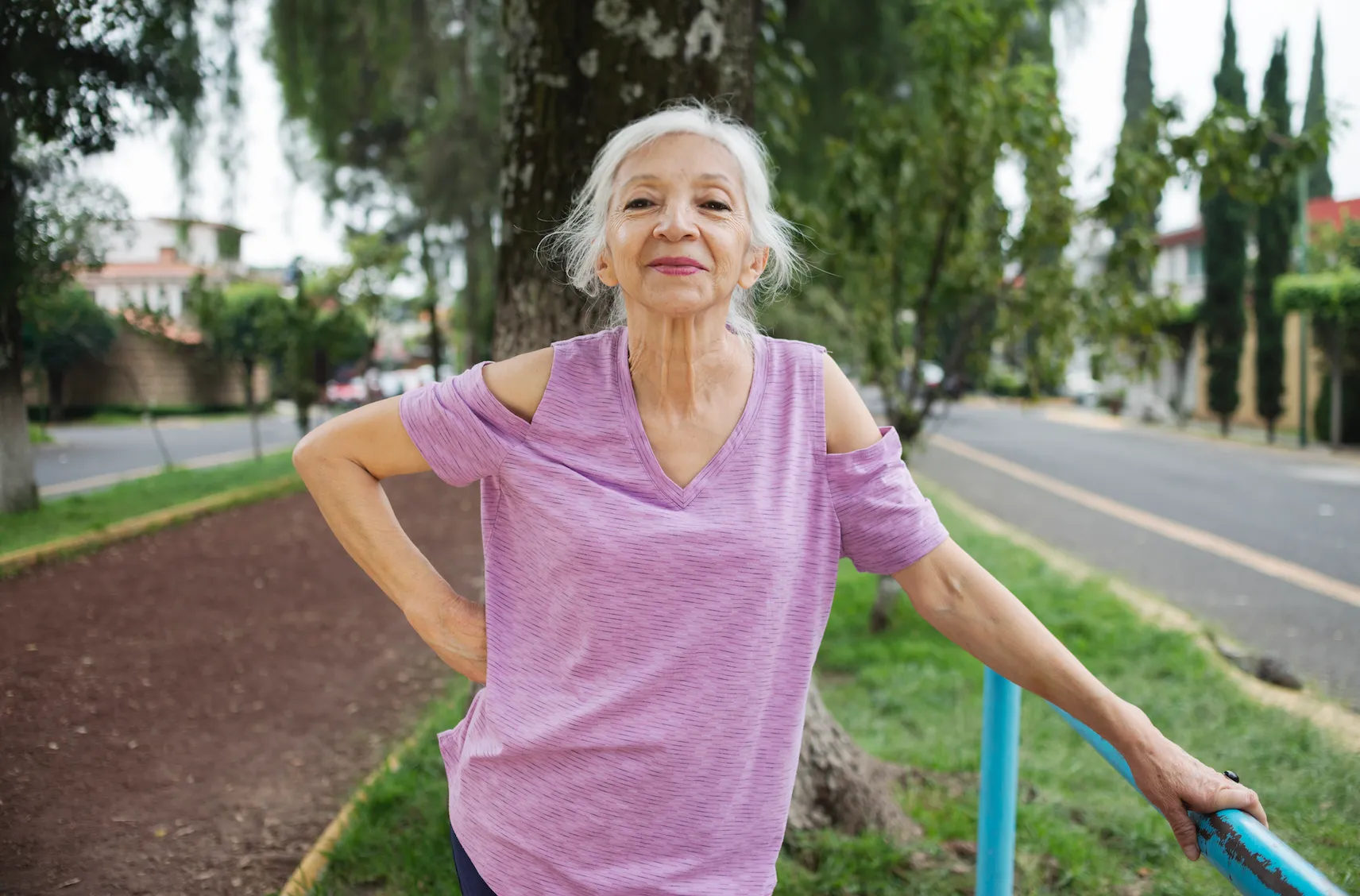 Senior woman in a purple shirt stretching outdoors near a park path and road.