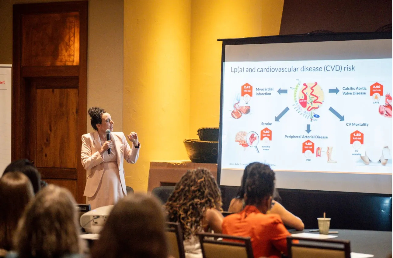 Woman in beige suit giving a presentation on cardiovascular disease risk to seated audience.