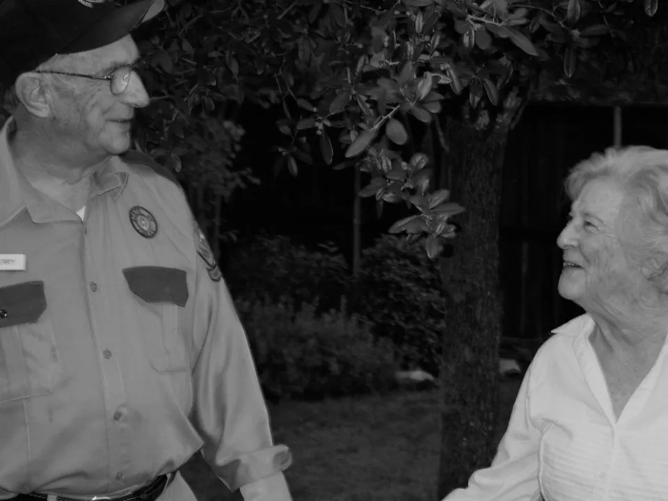 Black and white photo of an elderly man in uniform smiling at an elderly woman outdoors at night.